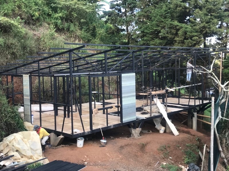 Estructura metálica negra de una cabaña OVO HOMES ensamblándose sobre una ladera arbolada, elevada sobre pilotes, con la placa de piso ya instalada y operarios trabajando
